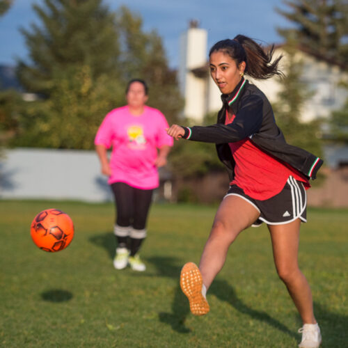 Woman in a red shirt and black shorts kicking an orange soccer ball.