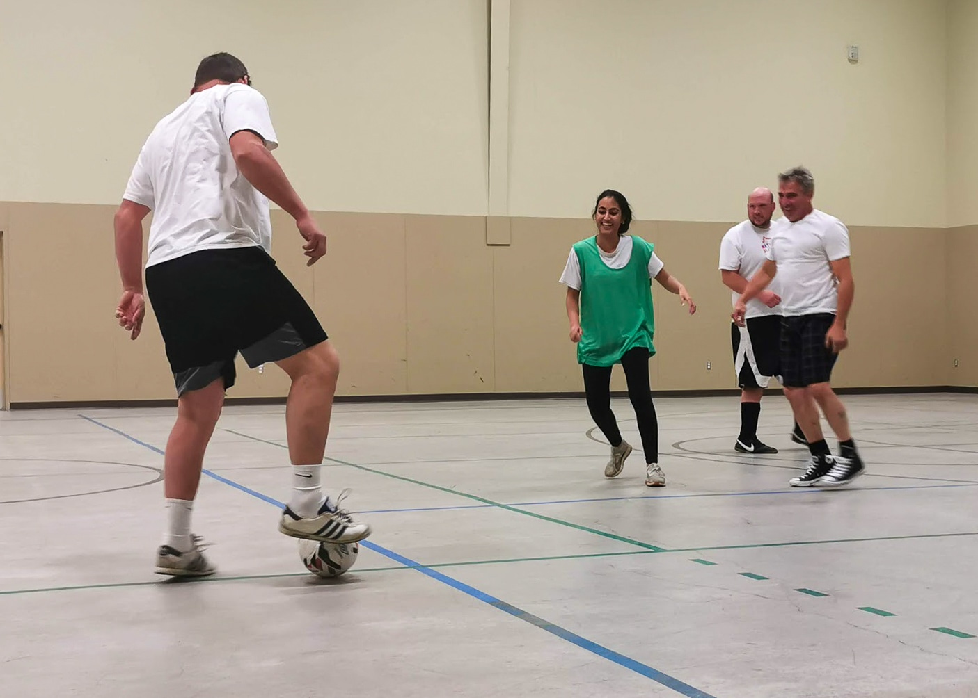 Adults playing soccer. Three men in white T-shirts and one woman wearing a green bib.